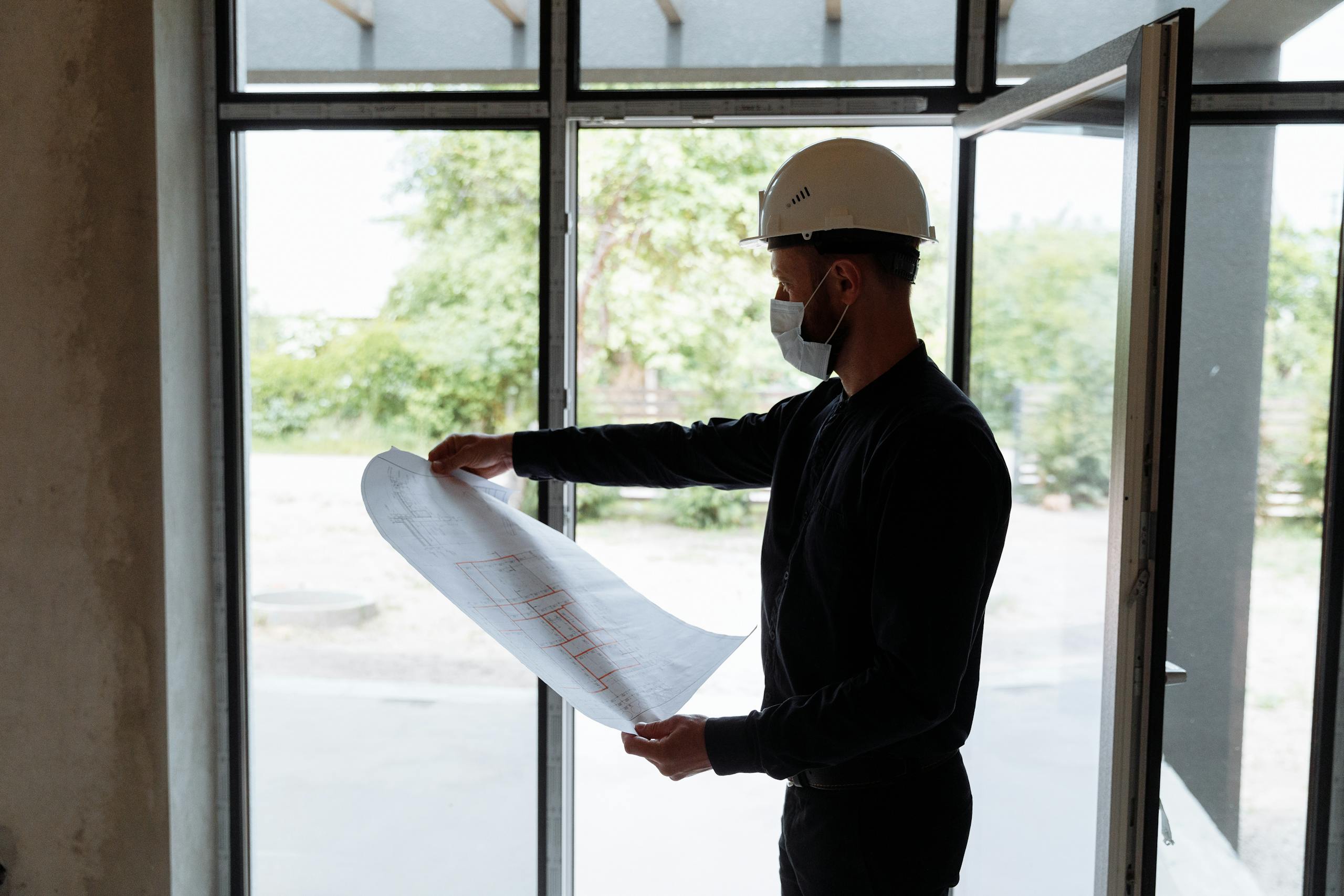 Architect reviewing floor plans indoors, wearing safety gear, focused on construction planning.